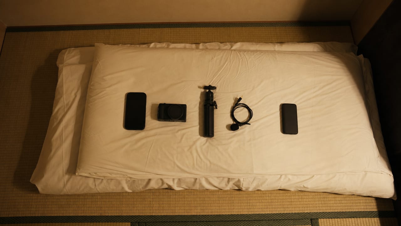 A whole filming kit laid out on a bed: a phone, a small camera, a folding tripod, a clip-on microphone and a battery